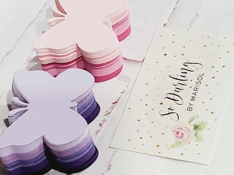 Puede incluir: Dos pilas de recortes de papel con forma de mariposa. La pila superior es rosa y la pila inferior es morada. Las pilas est&aacute;n sobre una superficie blanca. Una tarjeta blanca con purpurina dorada y el texto "So Darling by Marisol" est&aacute; en el fondo.