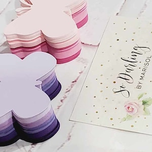 Puede incluir: Dos pilas de papel con forma de mariposa, una en tonos rosados y la otra en tonos morados. Las pilas est&aacute;n sobre una superficie blanca con una tarjeta de visita que dice "So Darling by Marisol" en letra cursiva negra.