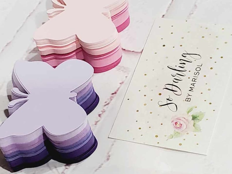Puede incluir: Dos pilas de papel con forma de mariposa, una en tonos rosados y la otra en tonos morados. Las pilas est&aacute;n sobre una superficie blanca con una tarjeta de visita que dice "So Darling by Marisol" en letra cursiva negra.
