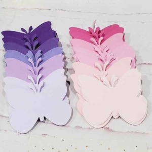 Puede incluir: Dos pilas de mariposas de papel, una en tonos morados y la otra en tonos rosados. Las mariposas est&aacute;n superpuestas de la m&aacute;s oscura a la m&aacute;s clara.