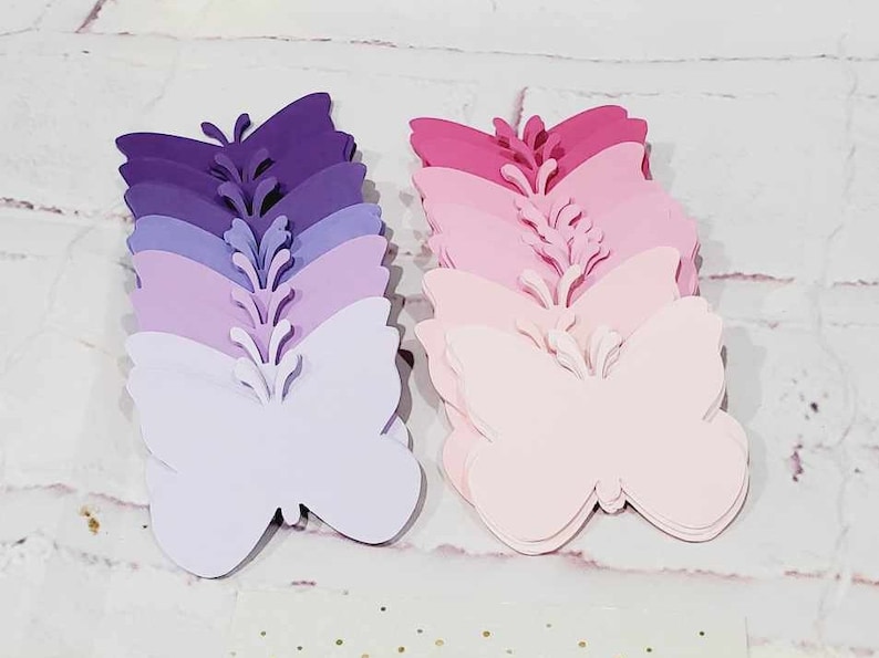 Puede incluir: Dos pilas de mariposas de papel, una en tonos morados y la otra en tonos rosados. Las mariposas est&aacute;n superpuestas de la m&aacute;s oscura a la m&aacute;s clara.