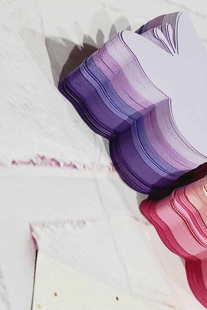 Puede incluir: Una pila de formas de mariposa de papel en capas en tonos de morado, rosa y blanco. Las mariposas est&aacute;n dispuestas en forma de coraz&oacute;n.