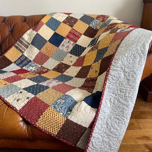 May include: A patchwork quilt featuring a grid of squares in various patterns and colours including deep red, navy blue, mustard yellow, and cream. The quilt has a wide, light grey border with a continuous swirling quilted stitch pattern throughout.