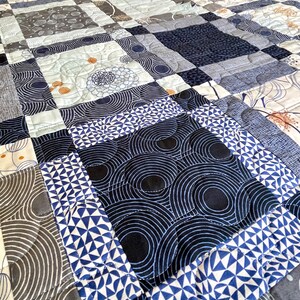 Op de afbeelding: Een quilt gemaakt van verschillende blauwe en grijze stoffen met patronen. De quilt is gemaakt van vierkanten en rechthoeken, en de patronen omvatten cirkels, geometrische vormen en bloemenmotieven.