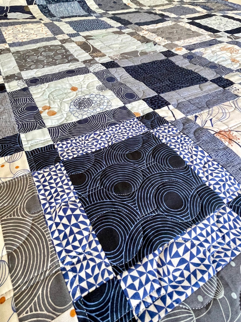 Op de afbeelding: Een quilt gemaakt van verschillende blauwe en grijze stoffen met patronen. De quilt is gemaakt van vierkanten en rechthoeken, en de patronen omvatten cirkels, geometrische vormen en bloemenmotieven.