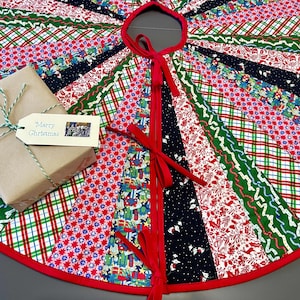 May include: A circular tree skirt featuring a patchwork of red, green, and navy fabric strips with holiday patterns. The skirt has a red fabric trim, red ribbon ties, and a brown paper gift box with a tag reading Merry Christmas.