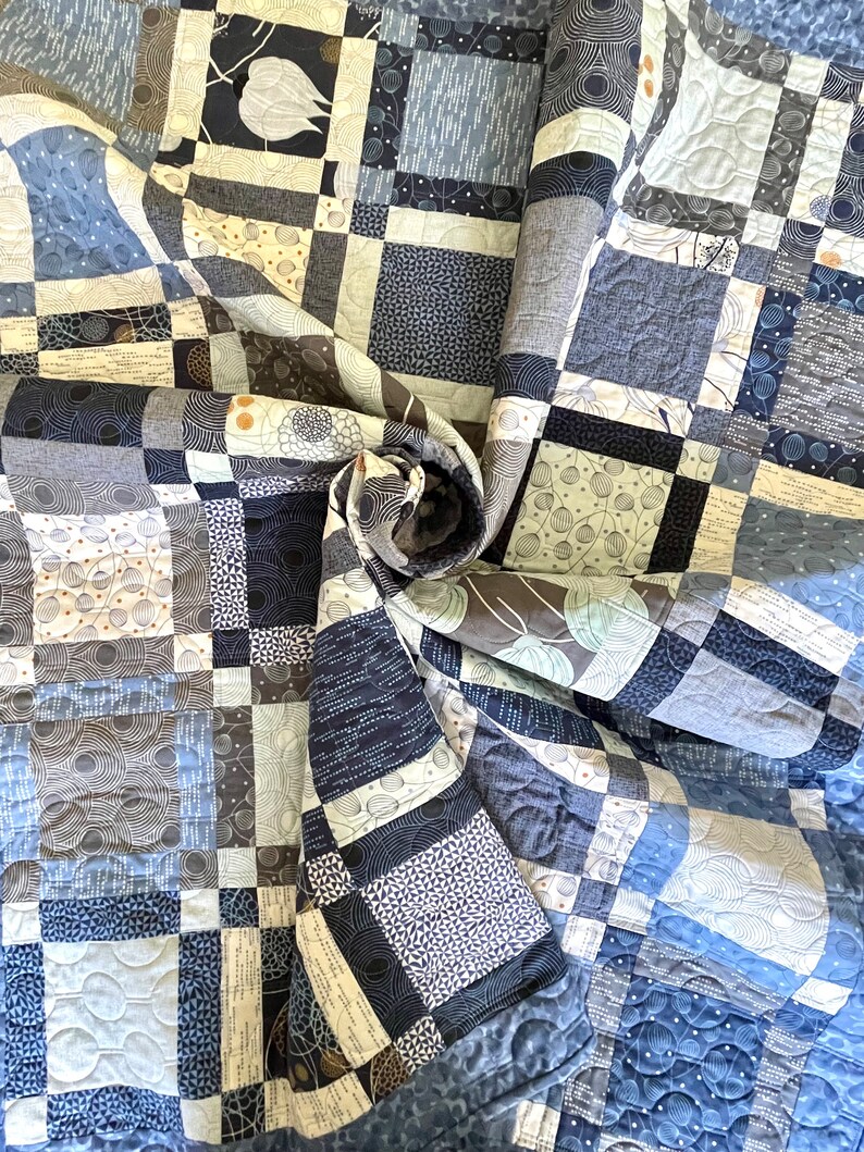 Op de afbeelding: Een blauwe en witte quilt met een geometrisch patroon. De quilt is gemaakt van verschillende stoffen met verschillende texturen en ontwerpen. De quilt is spiraalvormig gevouwen.