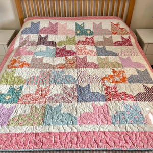 May include: A patchwork quilt with a repeating cat design. The quilt uses various patterned fabrics in pink, blue, green, and orange, set against a white backdrop. The quilt measures approximately 203 cm by 229 cm.