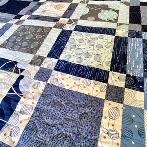 Op de afbeelding: Een quilt gemaakt van verschillende blauwe en witte stoffen met patronen. De quilt is gestikt met een golvend patroon. De quilt is gemaakt van vierkanten van verschillende groottes en patronen.