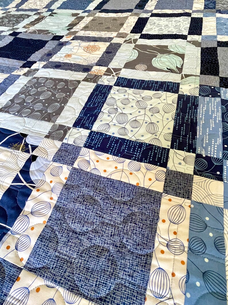 Op de afbeelding: Een quilt gemaakt van verschillende blauwe en witte stoffen met patronen. De quilt is gestikt met een golvend patroon. De quilt is gemaakt van vierkanten van verschillende groottes en patronen.