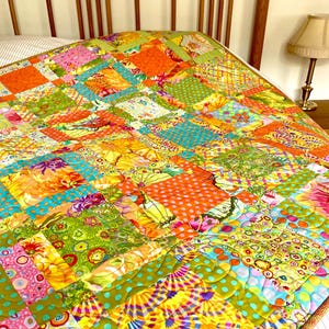 May include: A vibrant patchwork quilt featuring a mix of patterns and colours, including orange, green, pink, and blue. The quilt is constructed from squares and rectangles, creating a lively design. The quilt is displayed on a bed with a wooden headboard.
