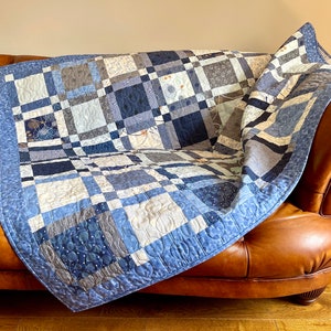 May include: A blue and grey patchwork quilt with a white background is folded on a brown leather sofa. The quilt is made of different fabrics with various patterns, including polka dots, stripes, and floral designs.