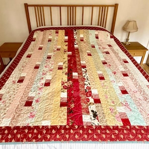 Puede incluir: Un edredón hecho a mano con un diseño de patchwork, con una variedad de telas florales y estampadas en tonos de rojo, rosa y crema. El edredón tiene un borde rojo con un patrón de hojas.