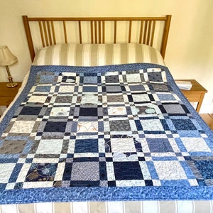 Op de afbeelding: Een blauwe en witte patchworkdeken met een lichtblauwe rand, getoond op een bed met een gestreept hoofdeinde en een wit en beige gestreept beddengoed.