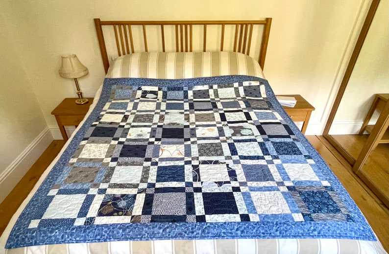 Op de afbeelding: Een blauwe en witte patchworkdeken met een lichtblauwe rand, getoond op een bed met een gestreept hoofdeinde en een wit en beige gestreept beddengoed.