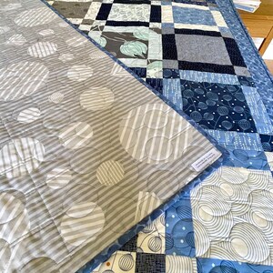 Op de afbeelding: Een blauwe en grijze quilt met een patchwork-ontwerp. De voorkant van de quilt heeft een geometrisch patroon met cirkels en vierkanten. De achterkant van de quilt heeft een patroon van cirkels met witte strepen. De quilt is gemaakt van katoenen stof en is machinewasbaar.