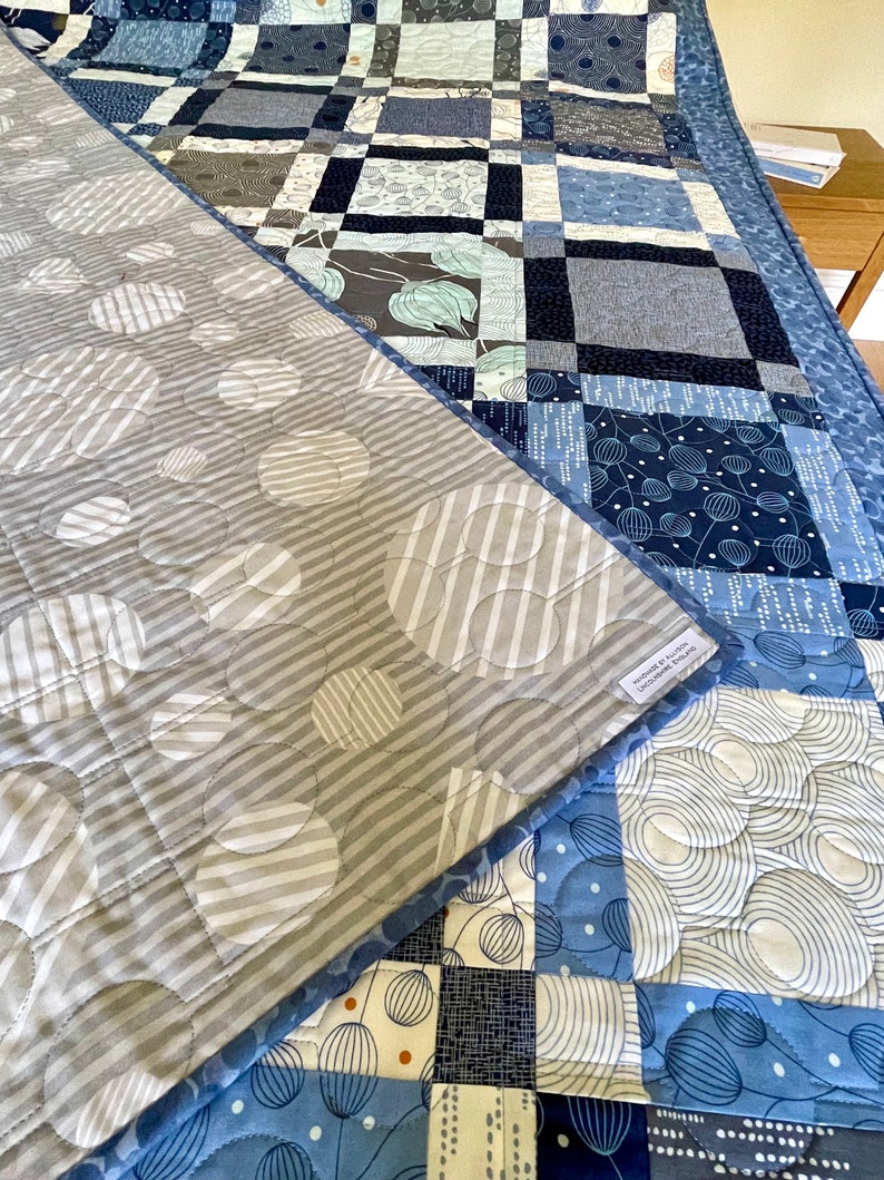 Op de afbeelding: Een blauwe en grijze quilt met een patchwork-ontwerp. De voorkant van de quilt heeft een geometrisch patroon met cirkels en vierkanten. De achterkant van de quilt heeft een patroon van cirkels met witte strepen. De quilt is gemaakt van katoenen stof en is machinewasbaar.