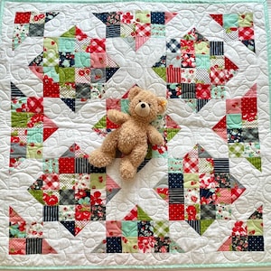 May include: A white quilted baby blanket with a patchwork design featuring red, green, blue, and pink floral patterns. A brown teddy bear is sitting in the center of the blanket.