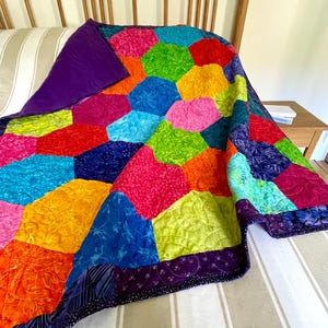 May include: A colorful quilt with a hexagonal pattern in shades of orange, yellow, green, blue, pink, and purple. The quilt is folded on a striped bedspread.