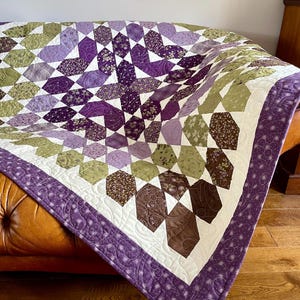 Könnte beinhalten: Handgefertigte Steppdecke mit geometrischem Muster in Lila-, Grün- und Brauntönen. Die Steppdecke ist über ein braunes Ledersofa drapiert. Der Rand ist einfarbig lila mit einem dekorativen Muster.