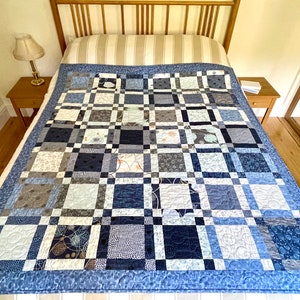 Op de afbeelding: Een blauwe en grijze patchworkdeken met een geometrisch patroon. De deken is gemaakt van verschillende stoffen met verschillende texturen en ontwerpen. Hij ligt op een bed met een houten hoofdeinde.