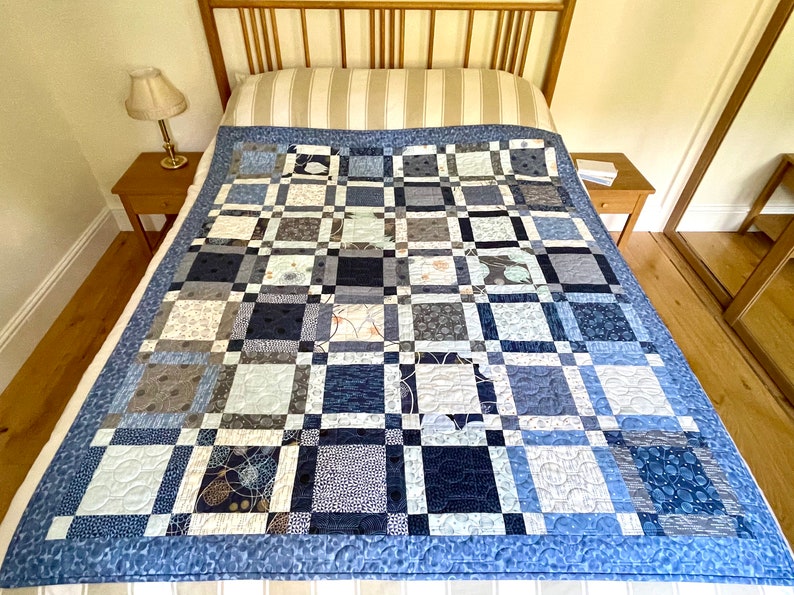 Op de afbeelding: Een blauwe en grijze patchworkdeken met een geometrisch patroon. De deken is gemaakt van verschillende stoffen met verschillende texturen en ontwerpen. Hij ligt op een bed met een houten hoofdeinde.