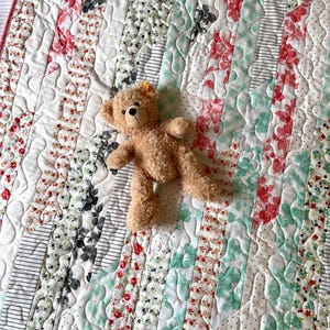 May include: A patchwork quilt with a variety of floral and striped patterns in shades of red, black, and green. A tan teddy bear is lying on top of the quilt. The quilt has a red border.