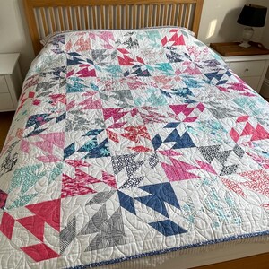 Könnte beinhalten: Eine handgefertigte Steppdecke mit einem geometrischen Muster in Rosa-, Blau- und Grautönen auf weißem Hintergrund. Die Steppdecke liegt auf einem Holzrahmen. Das Design der Steppdecke weist eine Vielzahl von Formen und Mustern auf.