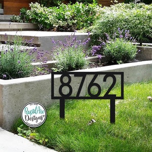 May include: Black metal house number sign with the numbers 8762 displayed in a white rectangle. The sign is on a green lawn in front of a concrete planter with lavender plants.
