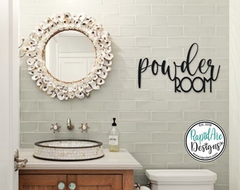 May include: Black metal sign with the words "powder room" in a cursive font. The sign is mounted on a white tiled wall in a bathroom. A white shell mirror is above a white tiled sink with a white countertop. A white toilet is visible in the background.