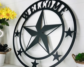 May include: A circular black metal wall sign featuring the word WELCOME cut out at the top. The design includes a large central five-pointed star surrounded by five smaller stars positioned along the lower inner edge of the circular frame.