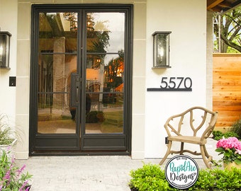 Puede incluir: Placa de número de casa de metal negro con el número 5570 en una pared blanca. La placa está montada sobre una puerta doble negra con paneles de vidrio. Hay un pequeño banco de madera y una planta en maceta con flores rosas frente a la casa.