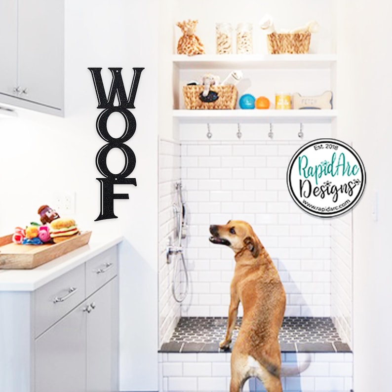May include: Black metal "WOOF" sign mounted on a white wall next to a dog shower stall. A brown dog stands in the shower stall, looking to the left. The shower stall has white subway tile walls and a black tiled floor. A logo for Rapid Arc Designs is visible in the upper right corner.