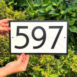 May include: A rectangular house number sign featuring the black digits 597 centered on a white background. The white surface has a subtle diagonal line pattern and is framed by a thick black border. Two small black mounting holes are visible.
