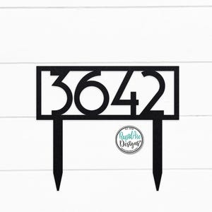May include: Black metal house number sign with the numbers 3642. The sign is on two black metal stakes. The sign is on a white wooden background.