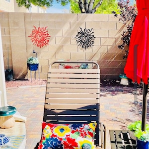 May include: An outdoor scene with a beige and black striped sun lounger and a floral cushion. A red parasol and small side table are to the right. Two metal sun wall hangings are on the brick wall behind the lounger.