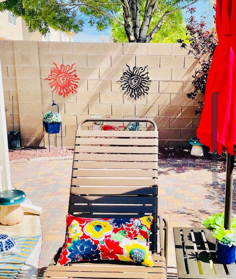 May include: An outdoor scene with a beige and black striped sun lounger and a floral cushion. A red parasol and small side table are to the right. Two metal sun wall hangings are on the brick wall behind the lounger.