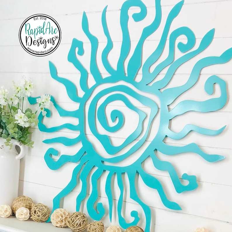 May include: A turquoise metal sunburst wall art piece with a layered design. The sun has a spiral centre and wavy rays. The Rapid Arc Designs logo is in the upper left corner. Decorative spheres and flowers are in the foreground.