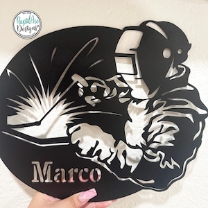 May include: A circular black metal wall art piece featuring a cutout silhouette of a welder wearing a helmet and holding a welding torch with sparks flying. The name Marco is cut out in bold letters at the bottom left of the circular metal sign.