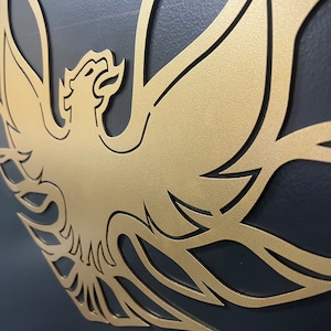 May include: A gold metal cutout of a stylized phoenix with intricate details. The phoenix is facing right and has its wings spread wide. The cutout is mounted on a dark gray background.