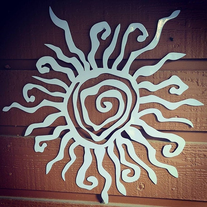May include: A white metal sunburst wall art piece. The sun has a spiral centre and wavy rays extending outwards. The artwork is mounted on a brown wooden surface, creating a contrast in colour and texture. This decorative piece is suitable for indoor or outdoor use.