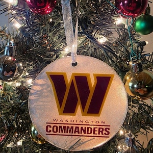 May include: A round, glittery Christmas ornament featuring the Washington Commanders logo in burgundy and gold. The ornament hangs from a silver ribbon and is surrounded by a Christmas tree with red, green, and gold ornaments.