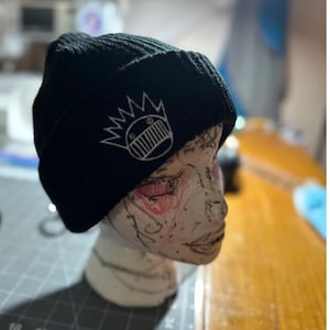 May include: Black knit beanie with a white embroidered graphic of a face with a crown on top.