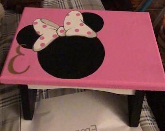 May include: A pink step stool featuring a black Minnie Mouse silhouette with a white bow and pink polka dots. The letter "E" is painted in gold. The stool has black legs.