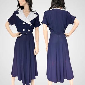 Könnte beinhalten: Ein marineblaues Vintage-Kleid mit weißem Kragen und weißen Knöpfen. Das Kleid hat kurze Ärmel und einen Gürtel in der Taille. Der Rock ist ein fließender Midirock. Das Kleid wird auf einer Schaufensterpuppe präsentiert.