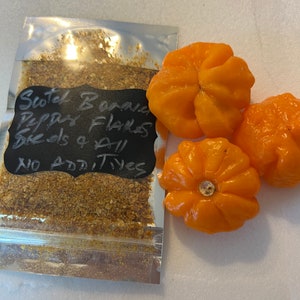 May include: A clear, sealed bag of Scotch Bonnet pepper flakes and seeds, with the text "Scotch Bonnet Pepper Flakes Seeds & All No Additives" on a black label. Three bright orange Scotch Bonnet peppers are next to the bag.