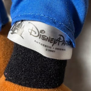 May include: Close-up of a Disney Parks label on a plush toy. The label is white with black text that reads "Disney Parks Authentic Original &copy; Disney". The toy has a blue shirt, black fur, and orange fabric.