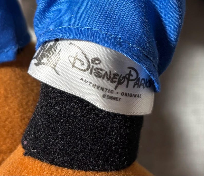 May include: Close-up of a Disney Parks label on a plush toy. The label is white with black text that reads "Disney Parks Authentic Original &copy; Disney". The toy has a blue shirt, black fur, and orange fabric.