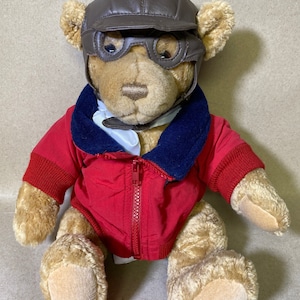 Puede incluir: Un oso de peluche marrón que lleva un casco de aviador de cuero marrón con gafas, una chaqueta roja con cuello azul y una camisa blanca.