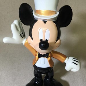 May include: A Mickey Mouse figurine in a formal outfit. The figure wears a white top hat with a gold band, a gold jacket, black pants, and black shoes. Mickey has white gloves and a black bow tie.